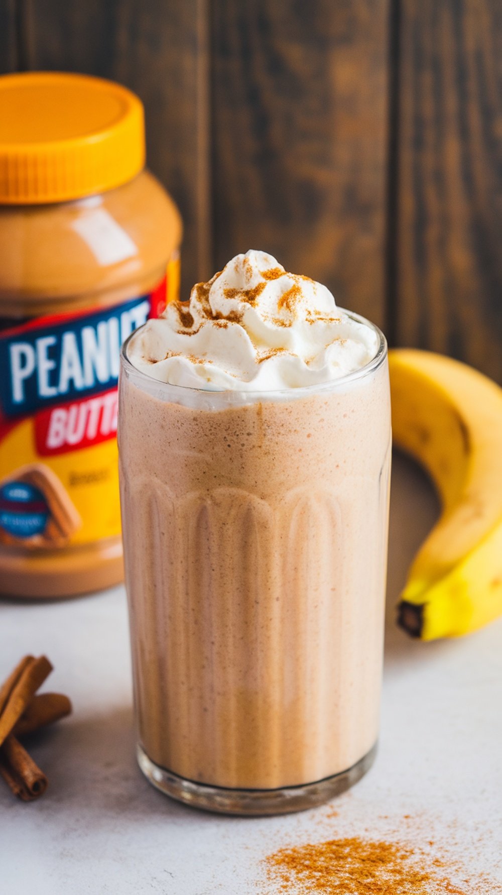A creamy peanut butter banana smoothie topped with whipped cream and cinnamon, with a jar of peanut butter and a banana in the background.